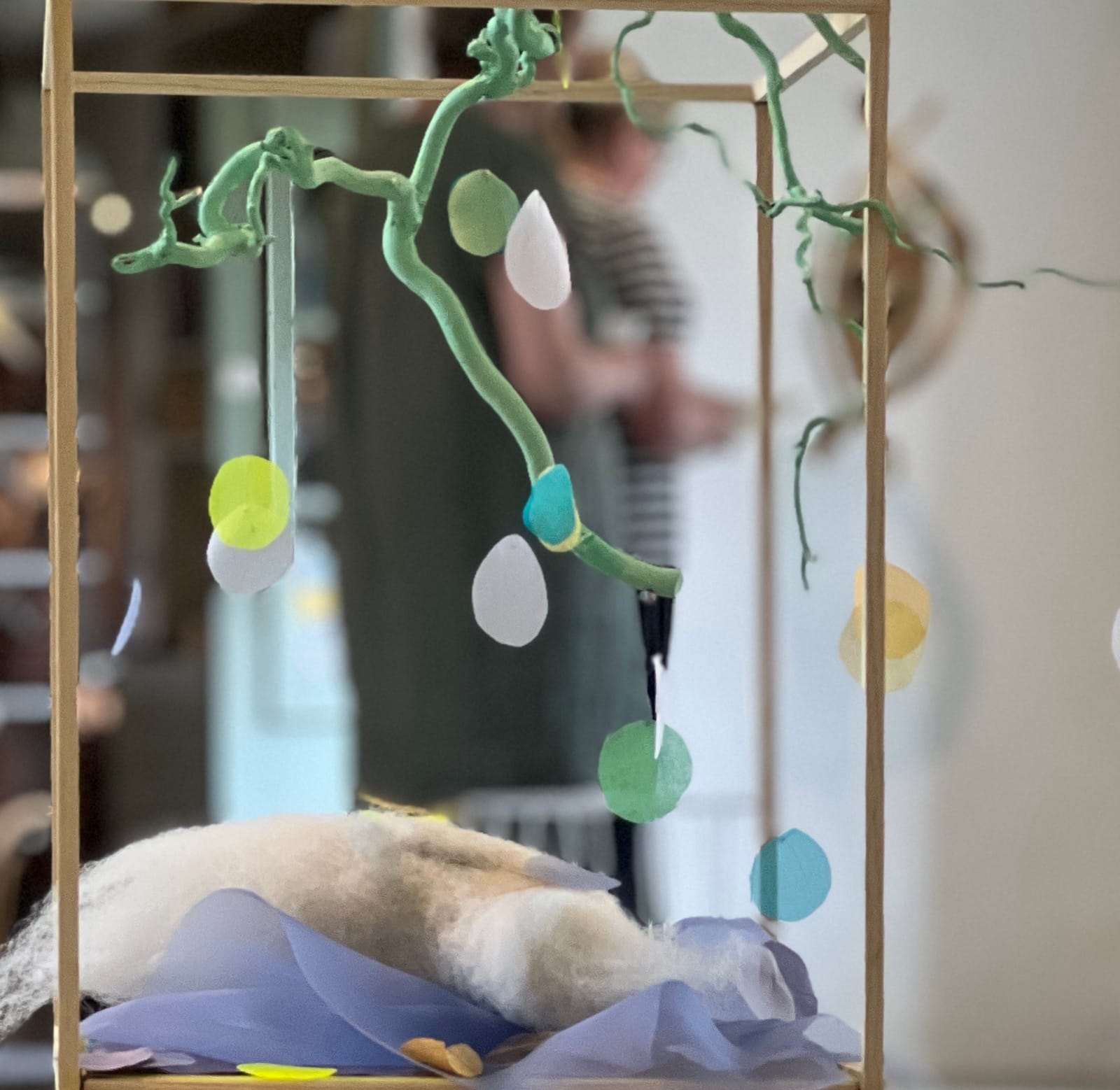 Blick genom kuben: den kittgröna grenen, löven i transparent tyg och en besökare som rör sig genom utställningsrummet i bakgrunden.