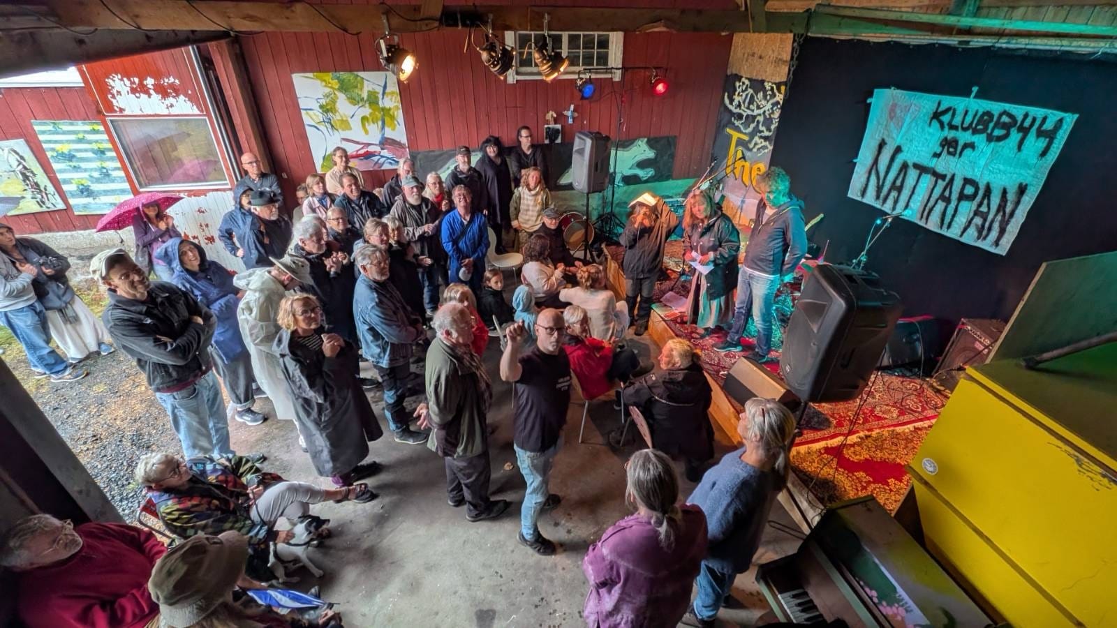 Festivaldagen 29 augusti: publik under takåsen framför målningar på den röda väggen, med scenen och KLUBB44-bannern i bakgrunden.