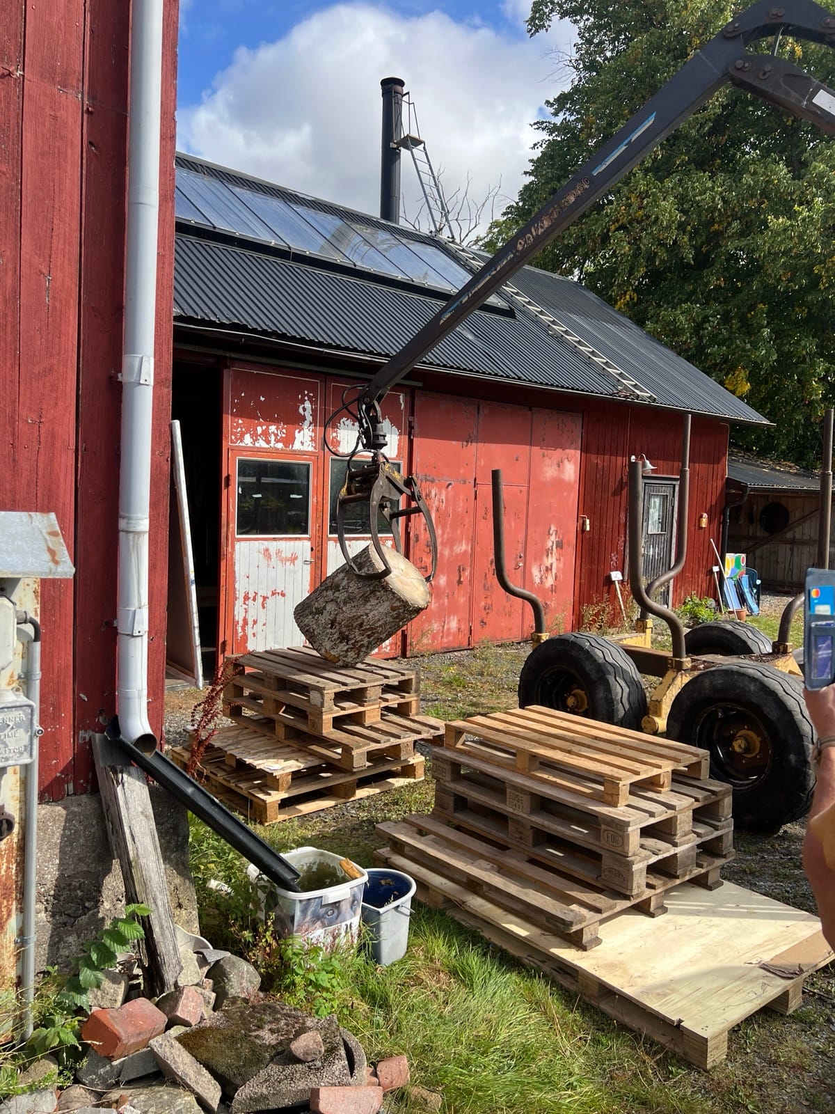 En stock lyfts av griplastaren och läggs på pallar framför den röda ladan – materialet till skulpturerna anländer.