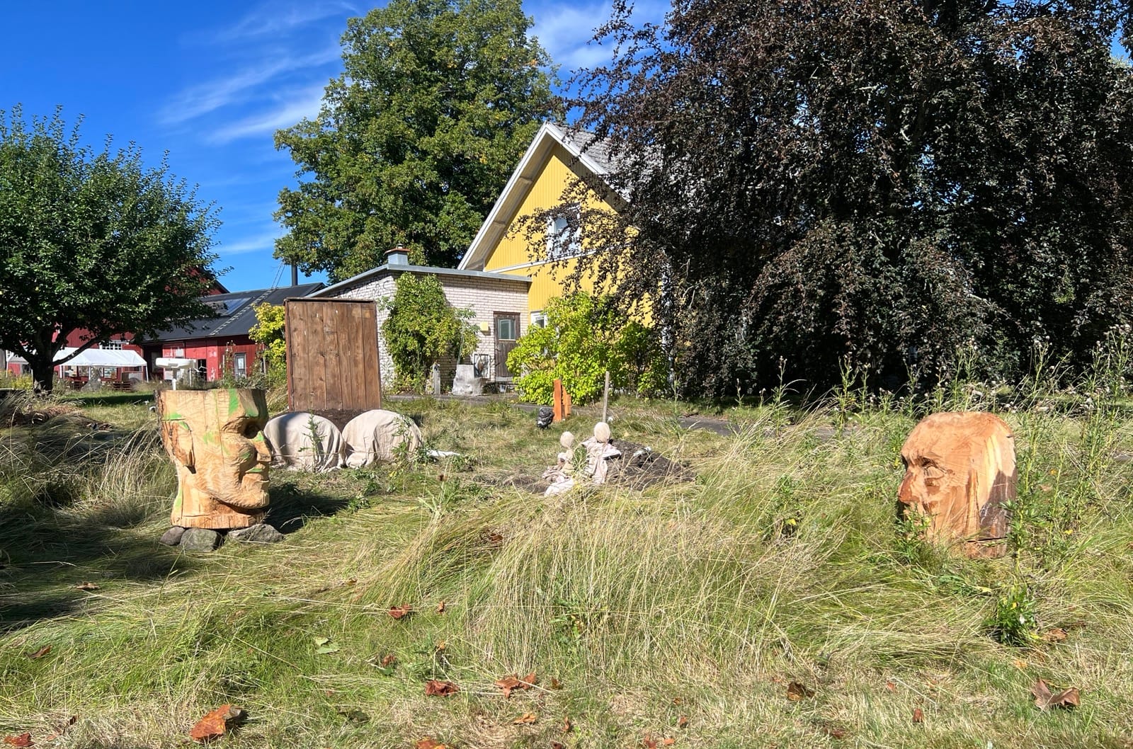 De två stora trähuvudena möts över ängen, med små gestalter och övertäckta former mellan sig – gården med det gula huset bakom.