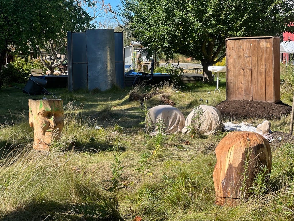 Trädgården med installationen: två stora trähuvuden, små övertäckta gestalter och mörka paneler bland fruktträd och högt gräs.