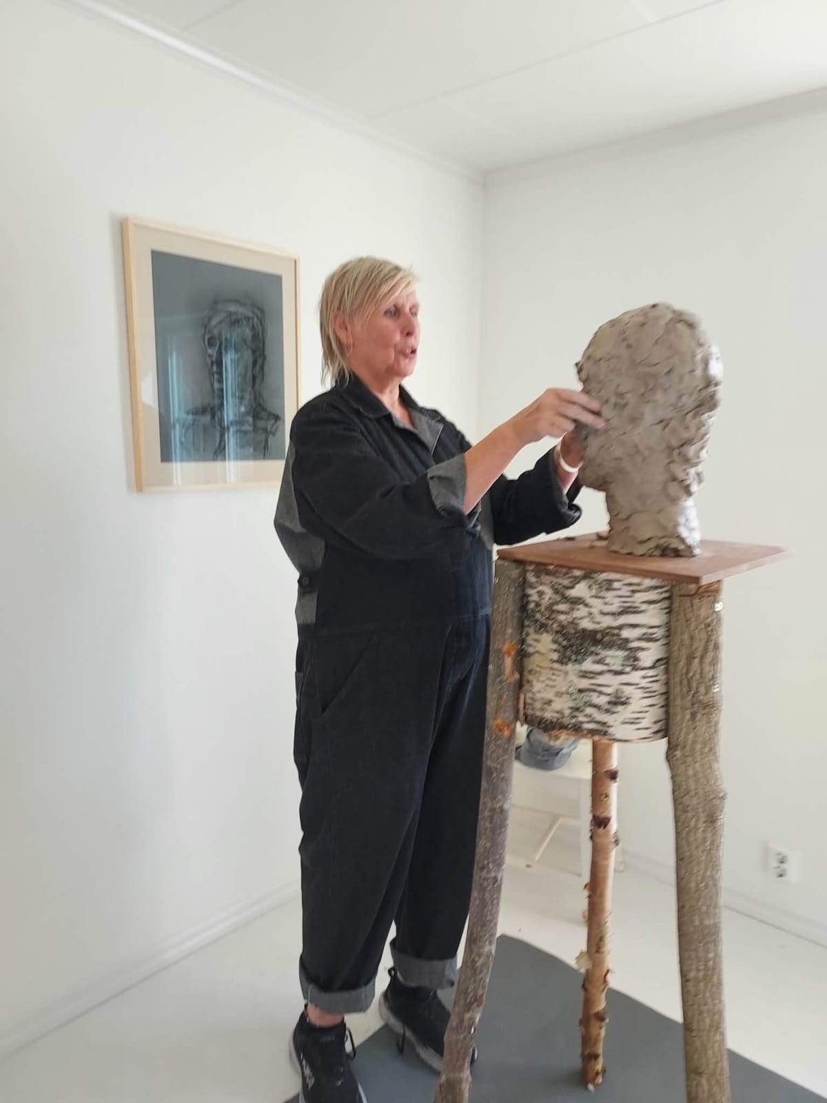 Lotte Nilsson Välimaa arbetar med en lerbyst i gallerirummet, med en inramad teckning på väggen bakom.