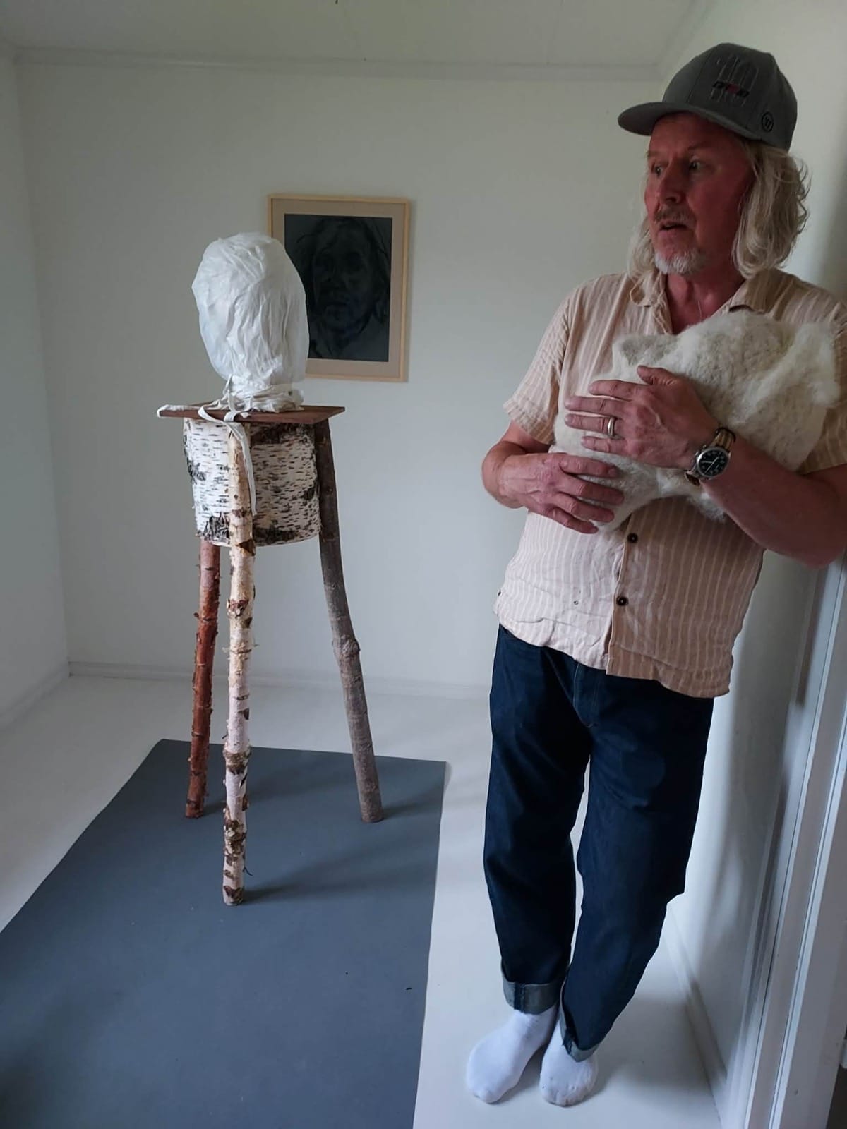 Kenneth Pils med en ullmassa i famnen, intill figuren med björkstammar och det inlindade huvudet i det vita gallerirummet.