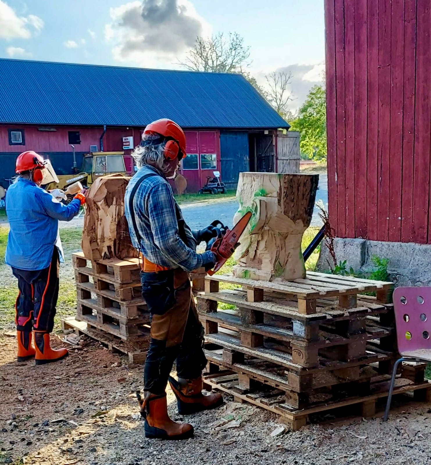 Lotte Nilsson Välimaa och Kenneth Pils arbetar med motorsåg på var sitt trähuvud, uppställda på pallar utomhus.